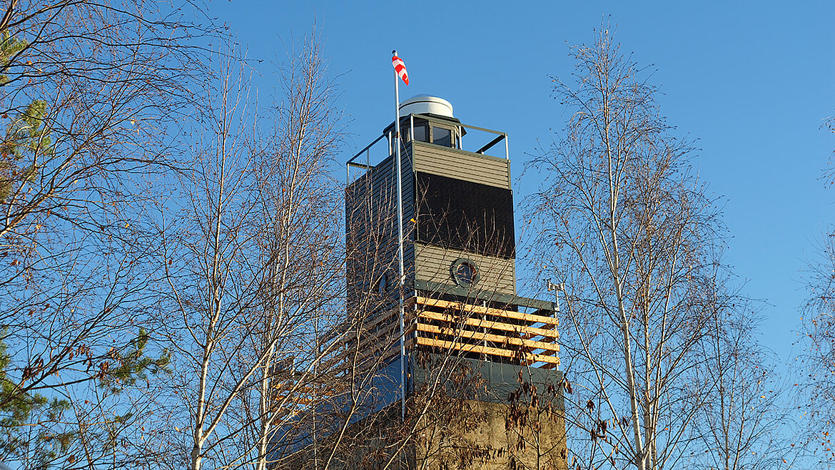 DIY-Leuchtturm mit Holzdetails und Fahne an der Spitze, umgeben von kahlen Bäumen vor blauem Himmel.
