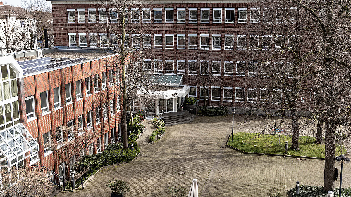 Außenansicht des Factory Campus Düsseldorf mit rotem Backsteingebäude, Glasdach und Innenhof.